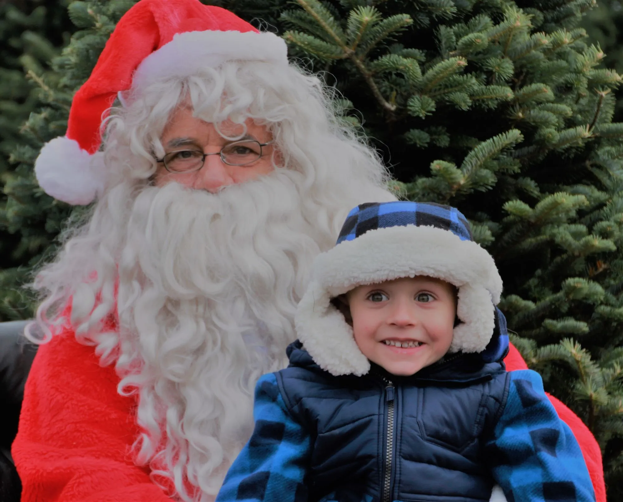 A boy in a fur-lined plaid hat grinning beside Santa at the Princeton Tree Farm